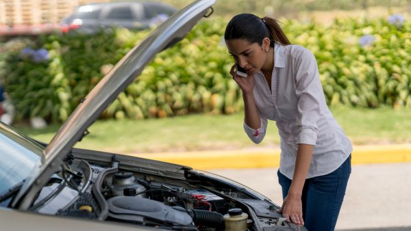 Mulher ligando para seguro do carro ao mesmo tempo que olha componentes do veículo após levantar capô