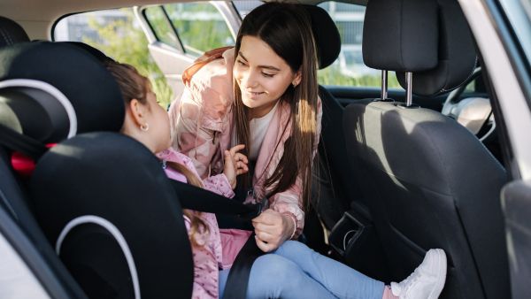 Mãe colocando cinto de segurança em filha no banco de trás de carro compacto